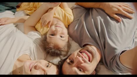 Close-up-shot-of-a-brunette-man-in-a-gray-T-shirt-taking-a-selfie-with-his-wife-a-brunette-girl-and-their-daughter-a-little-brunette-girl-in-a-yellow-dress-in-a-family-lying-on-the-sofa-and-taking-a-selfie-from-above-in-a-cozy-room