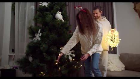 A-guy-in-a-white-sweatshirt-helps-his-girlfriend-hang-a-glowing-yellow-garland-on-the-New-Year-tree-in-a-cozy-home-in-winter.-Happy-couple,-a-guy-and-a-brunette-girl-are-preparing-for-the-New-Year-and-decorating-their-house