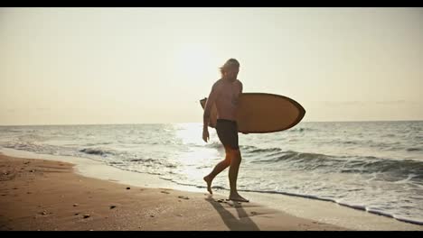 Ein-Blonder-Mann-Mit-Bart,-Nacktem-Oberkörper-Und-Grauen-Shorts-Trägt-Ein-Holzfarbenes-Surfbrett-Und-Geht-Morgens-Bei-Sonnenaufgang-Am-Sandstrand-Entlang