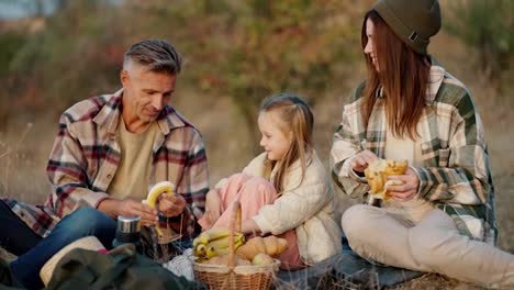 A-brunette-man-in-a-Green-checkered-shirt,-together-with-his-wife,-a-brunette-girl-in-a-Green-checkered-shirt-and-their-little-daughter,-are-sitting-outside-the-city-on-a-picnic-and-eating-fruits-in-the-summer-on-vacation