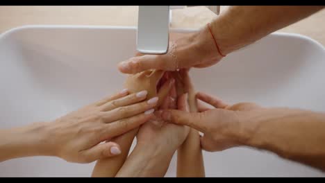 Close-up-shot-of-a-man-along-with-his-wife-and-daughter-washing-their-hands-in-a-modern-sink-under-running-water-against-the-background-of-a-white-porcelain-sink-in-the-bathroom