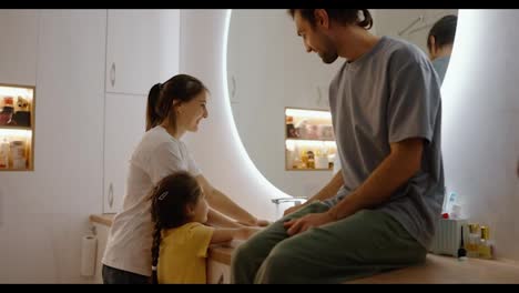 A-brunette-man-in-a-gray-T-shirt-sits-on-the-table-in-the-bathroom-and-looks-at-his-wife-and-daughter-washing-their-hands-together-in-a-modern-sink-near-the-mirror-in-a-modern-bathroom