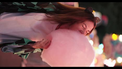 Vertical-video:-portrait-of-a-happy-brunette-girl-with-cat-paws-who-bites-and-tears-off-a-piece-of-large-pink-cotton-candy-during-her-walk-in-a-bright-amusement-park-in-the-evening