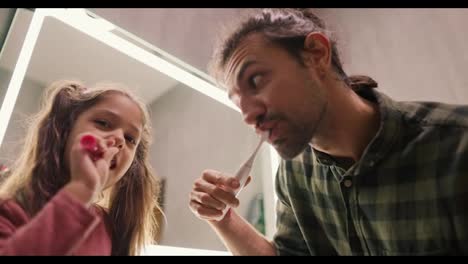 A-little-brunette-girl-with-a-ponytail-and-a-pink-dress-brushes-her-teeth-with-a-toothpick-along-with-her-single-father,-a-brunette-man-there-in-a-Green-checkered-shirt-near-a-mirror-in-a-modern-bathroom