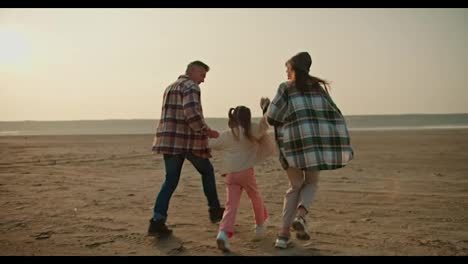 Rear-view-of-a-happy-brunette-girl-in-a-green-checkered-shirt,-together-with-her-husband,-a-middle-aged-man-with-gray-hair-in-a-checkered-shirt,-running-along-the-deserted-seashore-and-lifting-up-their-little-daughter,-a-blonde-girl-in-a-white-jacket-and-pink-pants,-during-their-vacation-outside-the