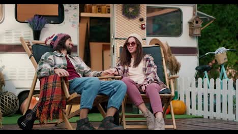 Happy-brunette-girl-in-round-glasses-and-a-white-hat-in-a-plaid-shirt-and-pink-pants-sits-on-a-chair-with-her-brunette-boyfriend-in-a-hat-near-a-trailer-during-their-picnic-at-a-camp-outside-the-city-in-the-summer