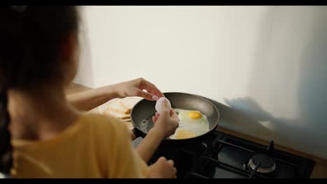 Rear-view-of-a-little-brunette-girl-in-a-yellow-dress-together-with-her-mother-preparing-fried-eggs-in-a-frying-pan-on-the-stove-in-a-modern-kitchen.-Girl-with-her-mom-learning-to-cook-breakfast,-family-morning-in-modern-apartment