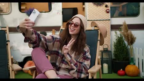 A-happy-girl-in-a-white-hat-and-plaid-shirt-sits-on-a-green-chair-against-the-backdrop-of-a-picnic-trailer-and-takes-a-selfie-using-a-white-smartphone-during-her-trip-outside-the-city-in-the-summer