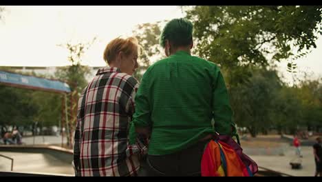 Rear-view-of-a-girl-with-green-hair-and-a-short-haircut-in-a-Green-shirt-and-a-blonde-girl-with-a-short-haircut-in-a-checkered-shirt-are-sitting-on-a-fence-in-a-skate-park-on-their-date-and-talking-to-each-other