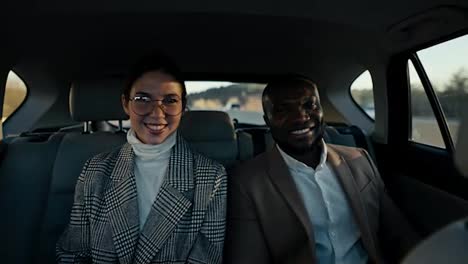 Portrait-of-a-happy-brunette-girl-in-round-glasses-a-businesswoman-in-a-gray-suit-and-her-male-colleague-with-Black-skin-in-a-brown-jacket-during-their-trip-in-a-modern-car-in-a-salon-outside-the-city