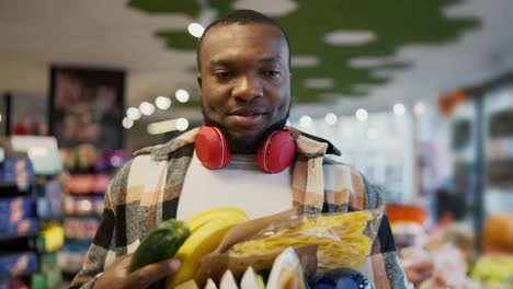 A-man-with-Black-skin-in-a-checkered-shirt-and-wearing-red-wireless-headphones-can-hardly-carry-a-large-amount-of-food-on-his-chest-with-both-hands.-The-man-does-not-use-a-bag-but-carries-a-lot-of-groceries-on-his-chest-holding-them-with-his-hand