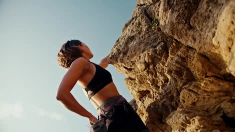 Shooting-from-below:-A-blonde-girl-in-a-black-top-and-black-pants,-using-a-special-harness,-climbs-a-rock-and-takes-sports-chalk-out-of-her-small-bag-for-more-confident-climbing-up-the-rock