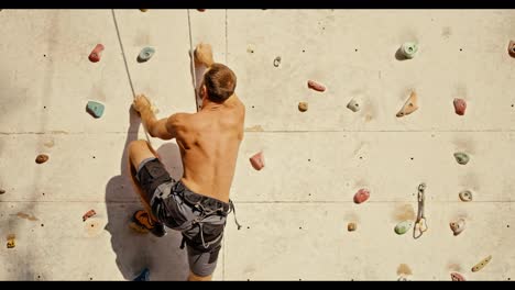 A-brunette-rock-climber-guy-in-gray-pants-with-a-sculpted-back-climbs-up-and-climbs-the-climbing-wall-on-a-sunny-summer-day.-Climber-guy-training-to-climb-rocks-using-a-training-structure-for-rock-climbers-in-the-summer