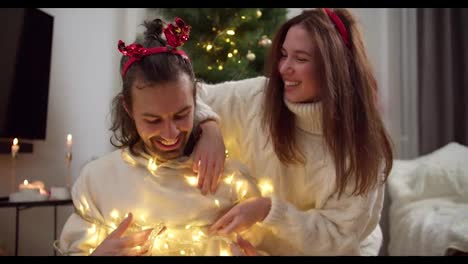 Close-up-shot-of-a-happy-brunette-girl-in-a-White-sweater-together-with-her-boyfriend-fooling-around-and-decorating-it-with-a-Christmas-glowing-yellow-garland-in-a-cozy-room-decorated-in-the-style-of-Christmas