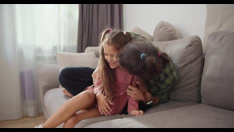 Happy-father,-a-brunette-man-in-a-green-checkered-shirt-plays-with-his-little-brunette-daughter-in-a-pink-dress-and-tickles-her-while-sitting-on-a-gray-sofa-in-a-modern-apartment