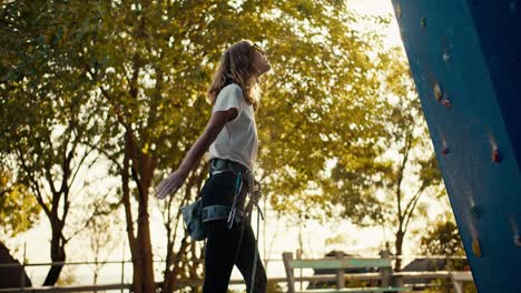 A-blonde-girl-climber-in-a-white-T-shirt-takes-out-sports-chalk,-rubs-it-and-begins-to-climb-up-the-blue-climbing-wall-on-a-sunny-summer-day