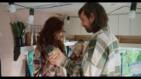 A-happy-brunette-guy-in-a-green-checkered-shirt-together-with-his-brunette-girlfriend-in-a-pink-checkered-shirt-choose-what-cereal-to-brew-for-breakfast-for-cooking-in-a-trailer-during-their-vacation-in-the-camp-in-the-summer