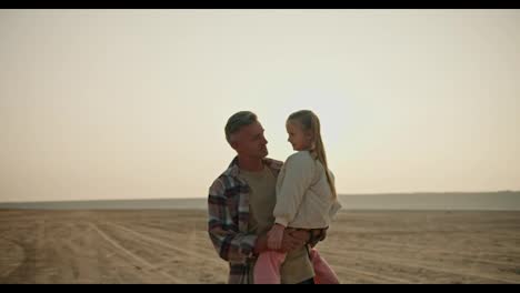 A-happy-middle-aged-brunette-man-with-gray-hair-in-a-checkered-shirt-walks-along-a-deserted-seashore-and-carries-in-his-arms-his-little-daughter,-a-blonde-girl-in-a-white-jacket,-during-his-hike-and-relaxation-outside-the-city-in-the-summer-evening