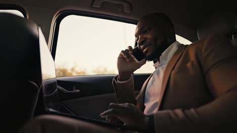 Happy-and-confident-man-Businessman-with-Black-skin-color-in-a-brown-jacket-communicates-on-a-smartphone-and-types-on-his-laptop-while-driving-In-the-passenger-seat-in-a-modern-car-outside-the-city