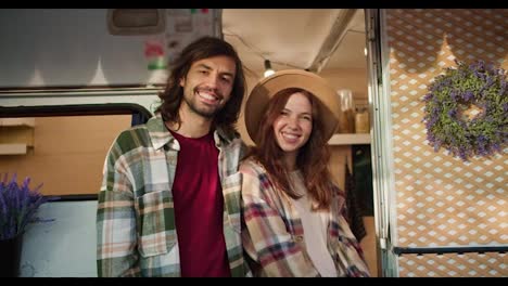 Portrait-of-a-happy-couple,-a-brunette-guy-with-stubble-in-a-Green-checkered-shirt-in-a-red-T-shirt-stands-near-his-brunette-girlfriend-in-a-brown-hat-and-checkered-shirt-Near-his-decorated-trailer-during-a-picnic-in-a-camp-outside-the-city-in-the-summer