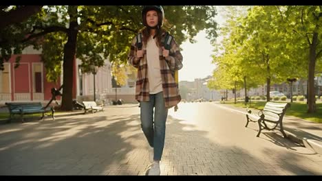 A-brunette-girl-in-a-checkered-shirt-and-denim-pants-with-a-white-motorcycle-helmet-approaches-her-victory,-sits-on-it-and-goes-to-work-as-a-courier