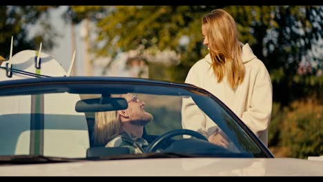 A-blonde-girl-in-a-white-sweatshirt-communicates-with-her-blond-boyfriend-with-a-beard-and-glasses-who-is-sitting-in-a-White-convertible-and-in-the-second-seat-he-has-surfboards-in-the-city-against-of-small-trees-with-green-foliage