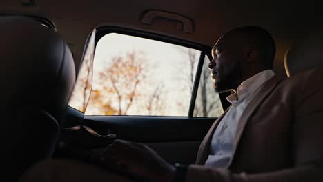 A-confident-man-with-Black-skin-and-a-beard-in-a-brown-jacket-works-on-a-laptop-and-looks-out-the-car-window-while-driving-a-modern-car-outside-the-city