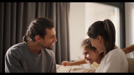 Close-up-shot-of-a-brunette-man-in-a-gray-T-shirt-and-his-wife,-a-brunette-girl-in-a-white-T-shirt,-hugging-their-little-daughter,-a-brunette-girl-in-a-yellow-dress-who-jumps-on-the-sofa-and-hugs-her-parents-in-a-cozy-modern-room