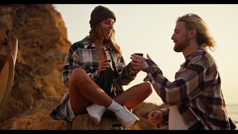 Happy-couple-blonde-girl-in-a-checkered-shirt-gives-her-boyfriend-a-mug-of-hot-tea-from-a-thermos-and-kisses-him.-A-blond-guy-with-a-beard-in-a-checkered-shirt-looks-at-his-girlfriend,-takes-tea-and-kisses-her-near-the-rocky-shore-in-the-morning-at-Sunrise-in-the