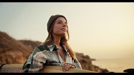 Close-up-shot-of-a-blonde-girl-in-a-green-hat-and-a-plaid-shirt-holding-a-yellow-surfboard-in-her-hands-standing-and-looking-at-the-sea-near-a-rocky-shore-by-the-sea-in-the-morning-at-sunrise