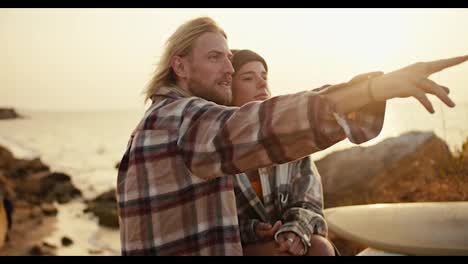 A-blond-man-with-a-beard-in-a-plaid-shirt-sits-next-to-his-blond-girlfriend-in-a-hat-and-a-plaid-shirt,-he-points-somewhere-to-the-side-and-they-laugh-on-the-rocky-earthen-shore-of-the-sea-at-Sunrise-in-the-summer,-their-surfboards-lie-near-them