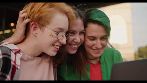 Close-up-shot-of-three-happy-girls-with-short-long-hair-in-bright-clothes-looking-at-something-on-a-laptop-screen-during-their-walk-in-the-summer