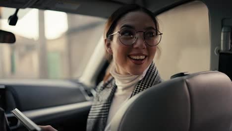 A-happy-brunette-businesswoman-girl-in-round-glasses-and-a-gray-jacket-communicates-with-her-interlocutor-who-is-sitting-in-the-back-seat-while-she-is-sitting-in-the-front-seat-in-a-modern-car-and-looking-at-something-on-her-tablet