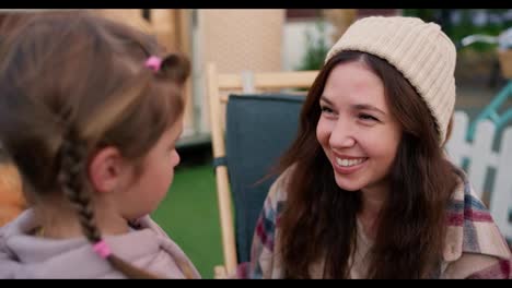 Close-up-shot-from-behind-the-shoulder-of-a-little-blonde-girl-in-a-pink-hoodie-talking-to-her-mother,-a-brunette-woman-who-hugs-the-girl-and-calms-her-down-during-their-picnic-near-a-trailer-in-a-summer-camp-outside-the-city