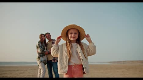 Portrait-of-a-happy-little-blonde-girl-in-a-white-jacket,-pink-pants-and-a-Straw-hat-who-stands-and-rests-during-her-picnic-with-her-parents,-a-brunette-man-with-gray-hair-and-a-brunette-girl-in-a-Green-checkered-shirt-on-a-deserted-seashore-in-summer