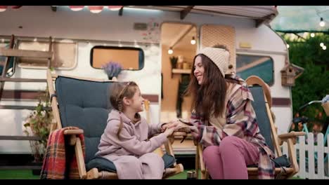 Happy-little-blonde-girl-in-a-pink-hoodie-communicates-with-her-mom,-a-brunette-woman-in-a-plaid-shirt-and-pink-pants,-during-her-vacation-in-a-camp-near-a-trailer-outside-the-city-in-summer