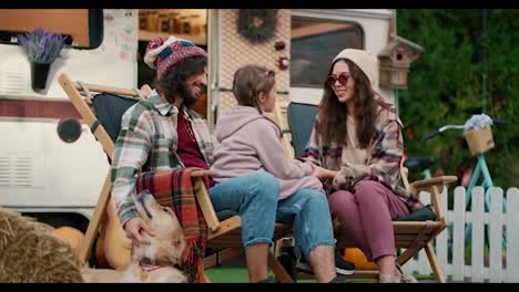 A-happy-brunette-guy-in-a-green-checkered-shirt-strokes-his-cream-colored-dog-and-his-girlfriend-communicates-with-his-little-daughter-who-sits-on-a-man-s-lap-near-a-trailer-in-the-camp-during-their-joint-vacation-and-picnic-outside-the-city-in-the-summer