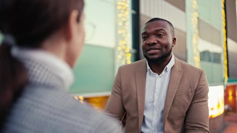 Over-the-shoulder-a-man-with-Black-skin-with-a-short-haircut-and-a-beard-in-a-brown-jacket-communicates-with-a-businesswoman-girl-in-round-glasses-and-a-gray-jacket-during-their-meeting-in-the-city