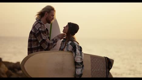 A-blond-guy-with-a-beard-in-a-plaid-shirt-straightens-his-hair-and-strokes-the-head-of-his-blonde-girlfriend-in-a-black-hat-and-plaid-shirt-who-is-holding-a-surfboard-standing-on-a-rocky-shore-near-the-sea-in-the-morning