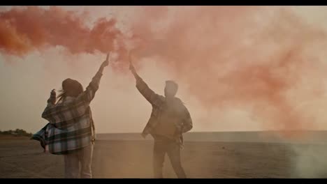 A-happy-brunette-man-with-gray-hair-in-a-checkered-shirt-together-with-his-wife,-a-brunette-girl-in-a-green-checkered-shirt,-holds-fireworks-in-their-hands-that-emit-green-and-red-smoke-during-their-relaxation-and-fun-outside-the-city-in-the-summer-evening