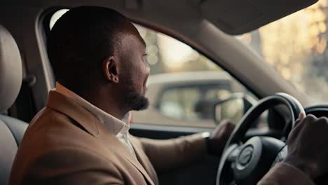 Un-Hombre-De-Negocios-Feliz-Y-Alegre-De-Piel-Negra-Y-Barba-Con-Una-Chaqueta-Marrón-Baila-Mientras-Está-Sentado-En-La-Cabina-De-Un-Automóvil-Moderno-Durante-Su-Viaje-De-Negocios-En-La-Ciudad.