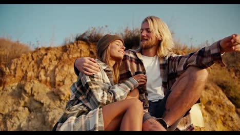 Happy-couple,-a-blond-man-with-a-beard-in-a-checkered-shirt-smokes-a-cigarette-while-sitting-with-his-blonde-girlfriend-in-a-checkered-shirt-and-a-green-hat-on-a-large-stone-on-a-rocky-shore-near-the-sea-and-points-somewhere-towards-the-sunrise-in-the-morning-near-their-paddle-boards-surf