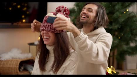 Happy-brunette-guy-puts-a-red-woolen-hat-on-his-brunette-girlfriend-in-a-white-sweater-hugs-her-and-rejoices-in-a-cozy-room-decorated-in-Christmas-style-with-a-Christmas-tree