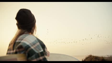 Rear-view-of-a-blonde-girl-in-a-Green-hat-in-a-plaid-shirt-stands-near-the-sea-with-a-surfboard-in-her-hands-and-looks-at-a-large-flock-of-birds-that-fly-somewhere-in-the-fall