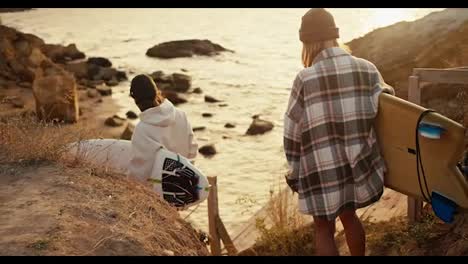 A-brunette-man-in-a-white-sweatshirt-and-a-blonde-woman-in-a-plaid-shirt-are-carrying-surfboards-while-going-down-a-wooden-staircase-to-a-rocky-shore-near-the-sea-at-sunrise-in-summer-in-the-summer