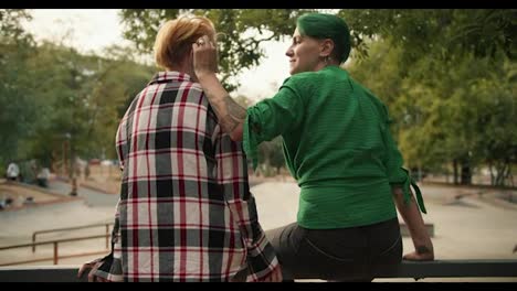 Rear-view-of-a-girl-with-short-green-hair-in-a-green-shirt-straightens-the-hair-of-her-girlfriend-with-yellow-hair-in-a-plaid-shirt.-Lesbian-couple-on-a-date-at-the-skate-park