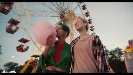 A-blonde-girl-with-a-short-haircut-in-a-pink-plaid-shirt-and-a-girl-with-green-short-hair-in-a-green-shirt-holds-a-huge-pink-cotton-candy-near-the-ferris-wheel-in-the-amusement-park-during-their-date