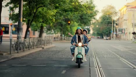 Un-Chico-Feliz-Con-Una-Chaqueta-Vaquera-Viaja-Con-Su-Novia,-Que-Mueve-Los-Brazos-Y-Baila-Mientras-Conduce-Un-Ciclomotor-Con-Las-Luces-Encendidas-En-Una-Calle-De-La-Ciudad-Por-La-Mañana-En-Verano.