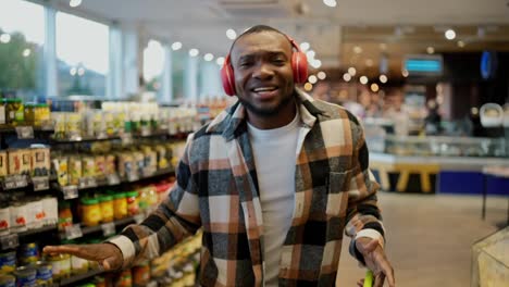 Portrait-of-a-happy-man-with-Black-skin-color-in-a-plaid-shirt-and-red-wireless-headphones-dancing-while-in-a-large-modern-grocery-store.-Happy-man-dancing-during-his-shopping-trip-and-shopping-at-the-grocery-store