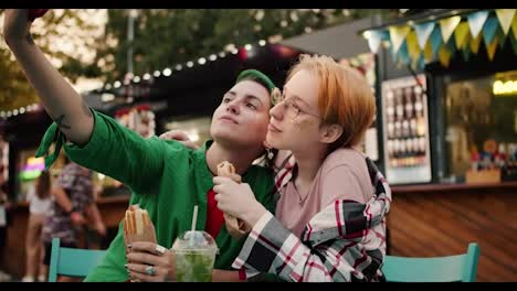 Una-Chica-De-Pelo-Corto-Verde-Con-Una-Camisa-Verde-Se-Toma-Una-Selfie-Con-Su-Novia,-Una-Rubia-De-Pelo-Corto-Con-Gafas-Y-Una-Camisa-A-Cuadros,-Usando-Un-Teléfono-Rojo.-Cuándo-Ir-De-Un-Hot-Dog-Cerca-De-Un-Café-Callejero-En-Verano-Al-Parque